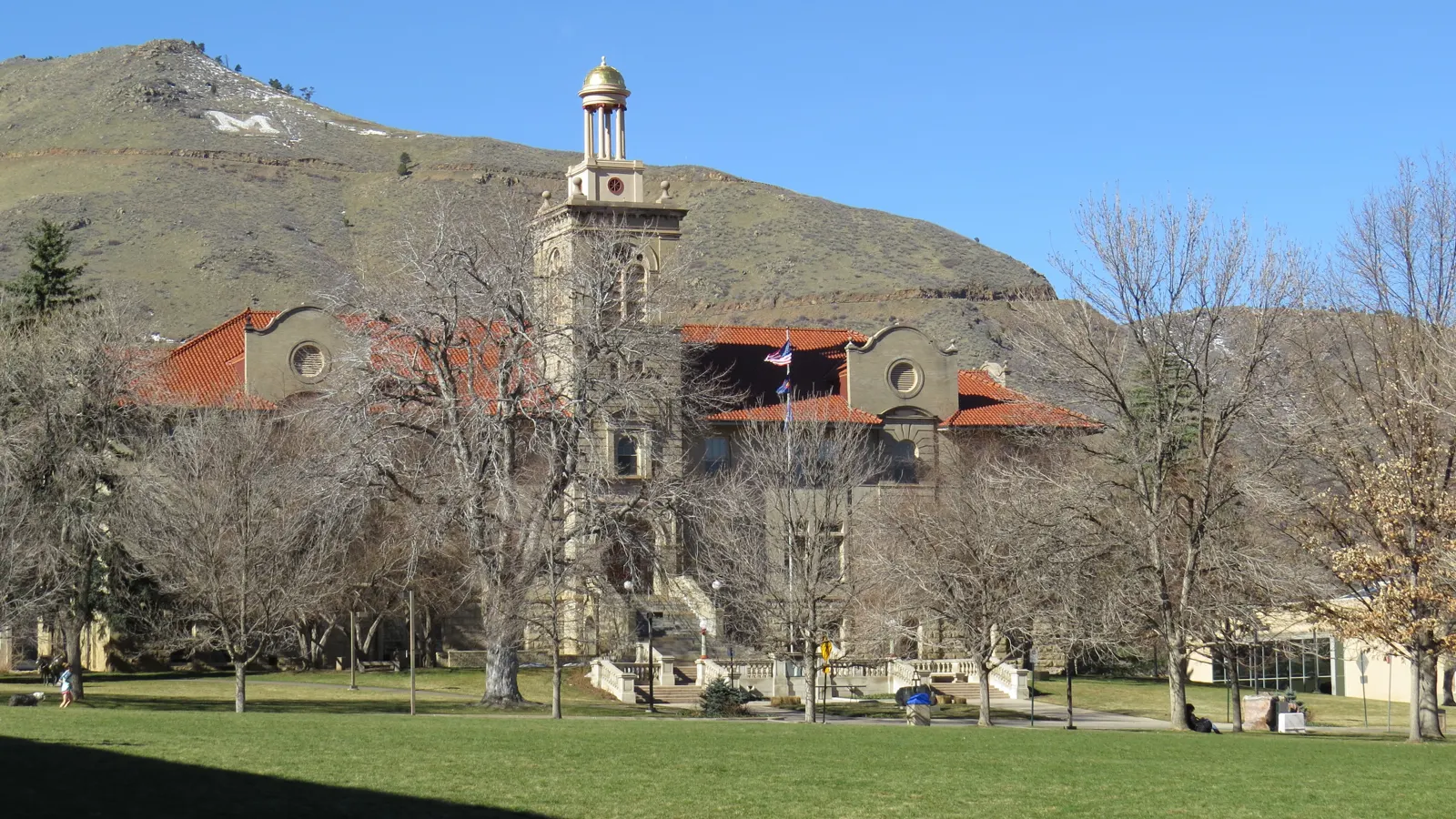 Guggenheim Hall at Colorado School of Mines, with its clock tower and red tile roof, across a lawn below the hillside white M in Golden, Colorado