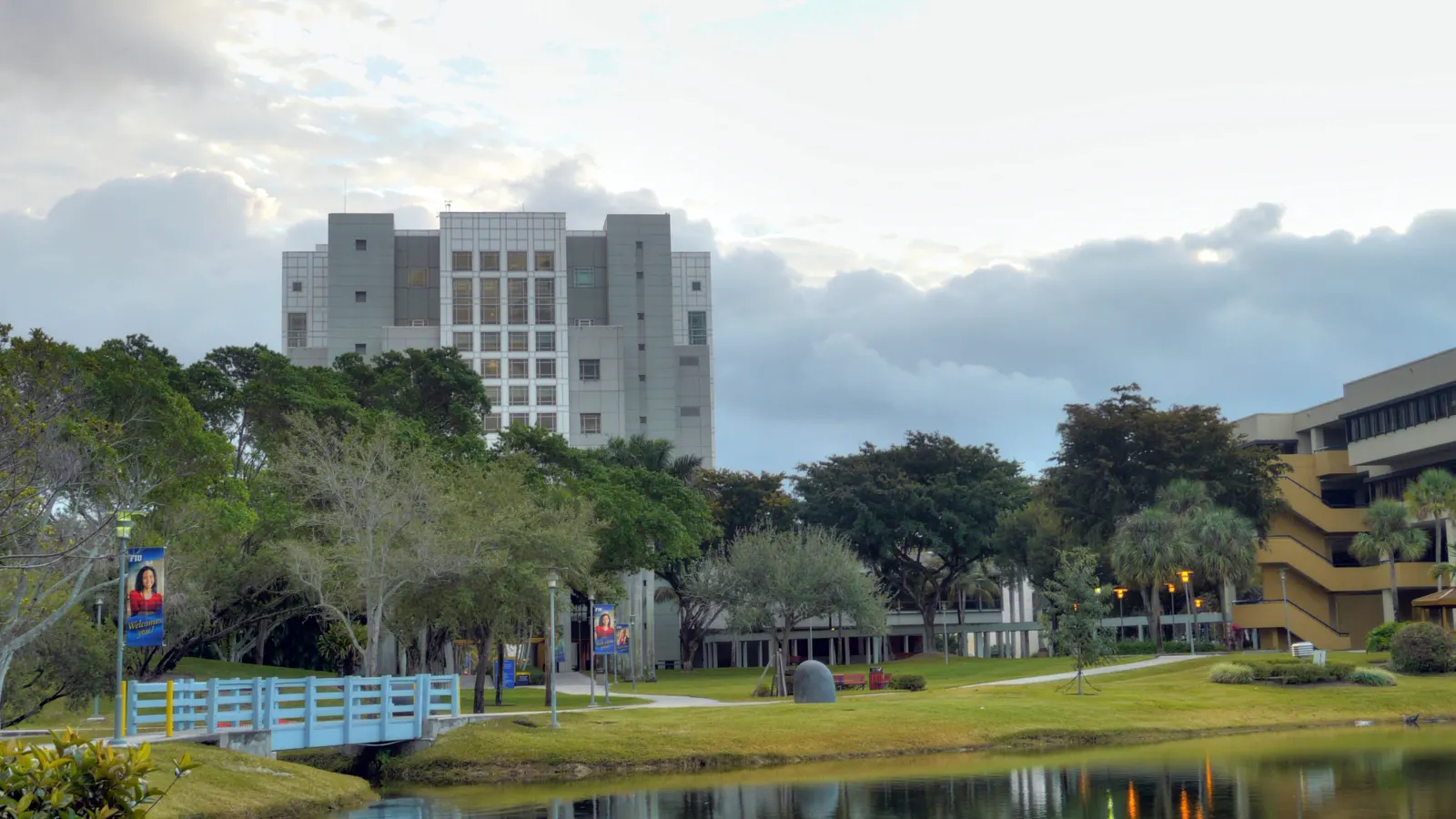 A lake and footbridge on the Florida International University campus in Miami, with campus buildings rising behind the trees
