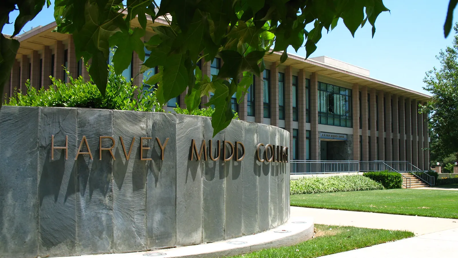 A curved stone wall with the Harvey Mudd College name in front of a campus building on a sunny day