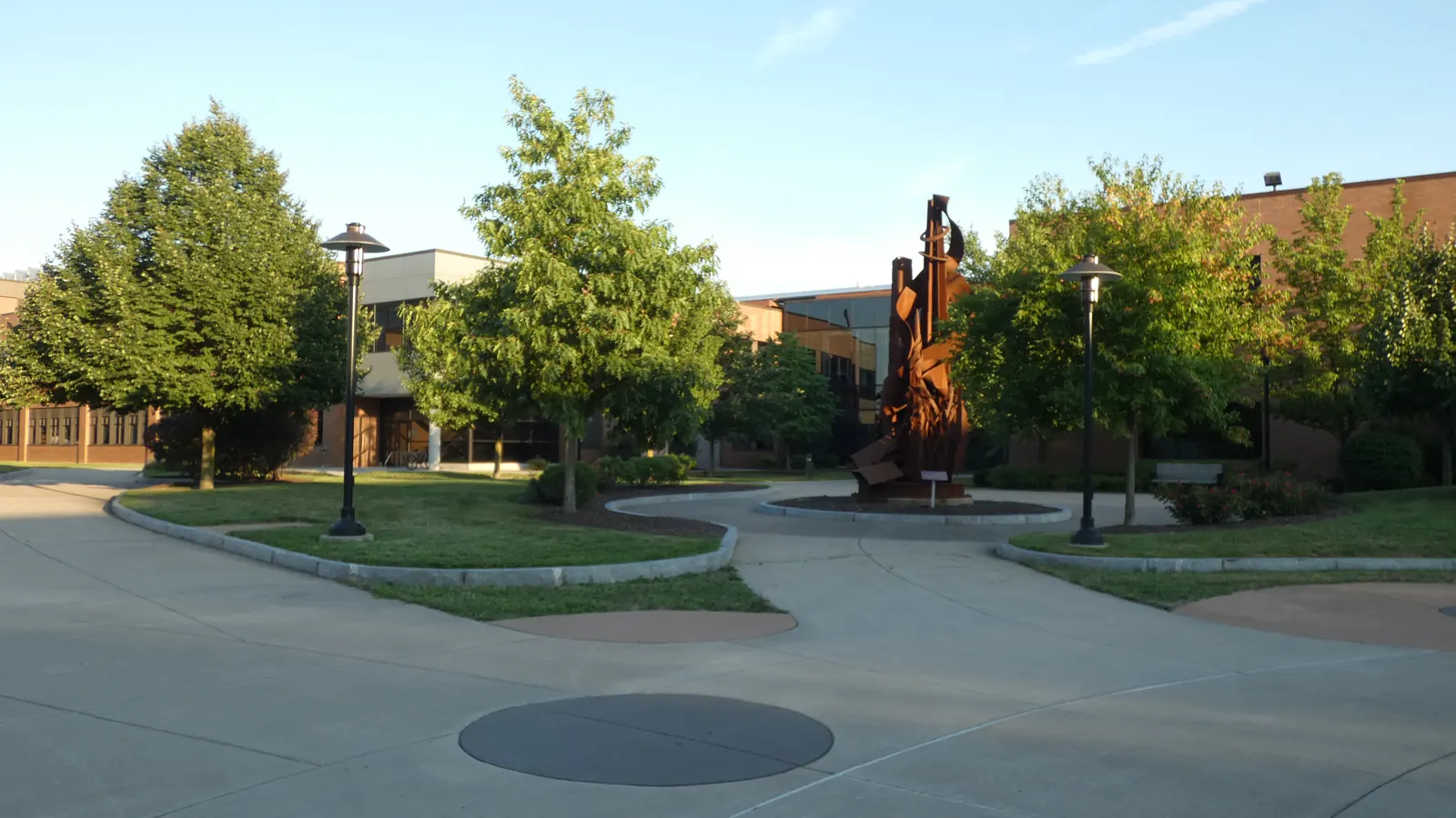 Brick campus buildings, trees and a tall rust-colored steel sculpture on a paved plaza at Rochester Institute of Technology