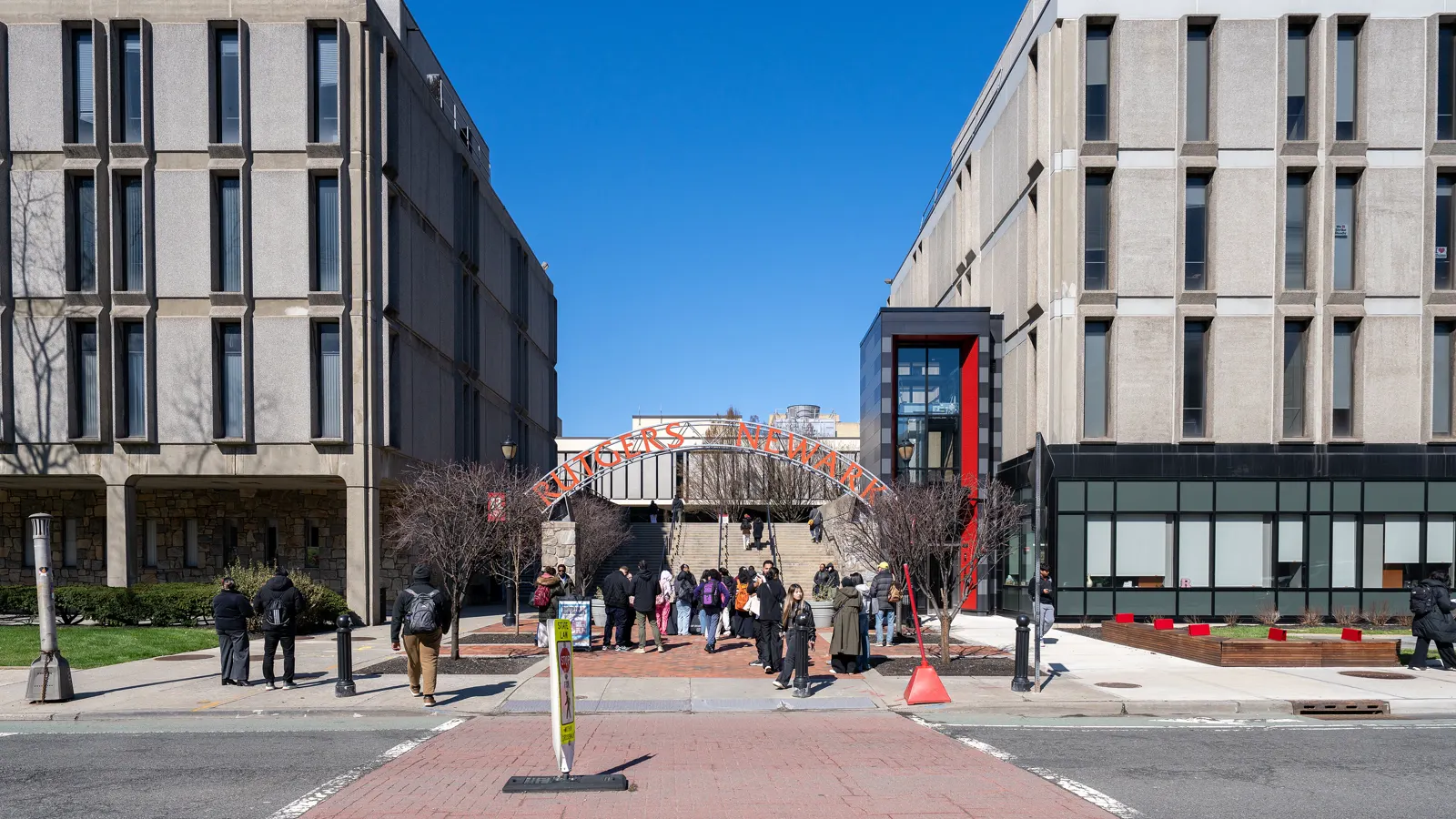 Students walk under the red Rutgers Newark arch between two campus buildings on a sunny day