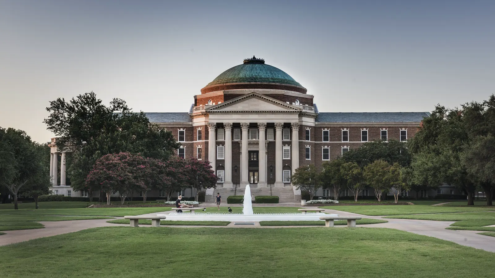 Dallas Hall at Southern Methodist University, a red brick building with white columns and a green copper dome, behind a lawn and a fountain at dusk