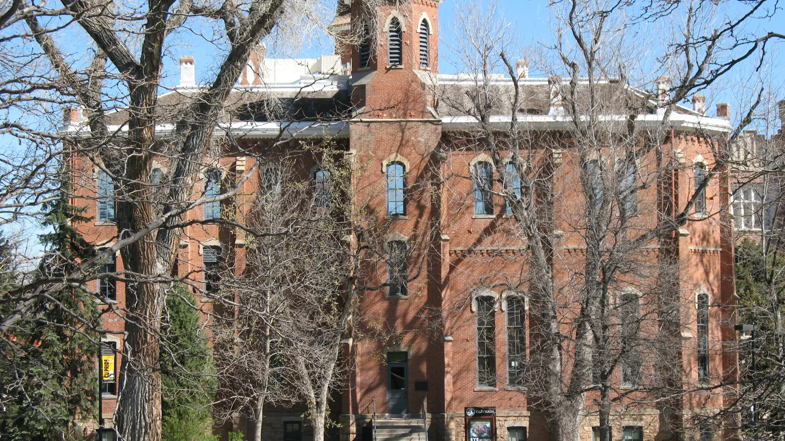 Old Main, a red brick building with a central tower, behind bare trees at the University of Colorado Boulder
