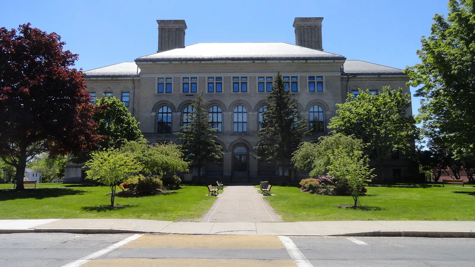 Coburn Hall, a stone building with arched windows and twin chimneys behind a tree-lined lawn, on the South Campus of the University of Massachusetts Lowell